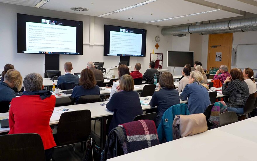 Teilnehmende sitzen im Hörsaal in Reihen an Tischen mit Blick auf zwei Bildschirme.