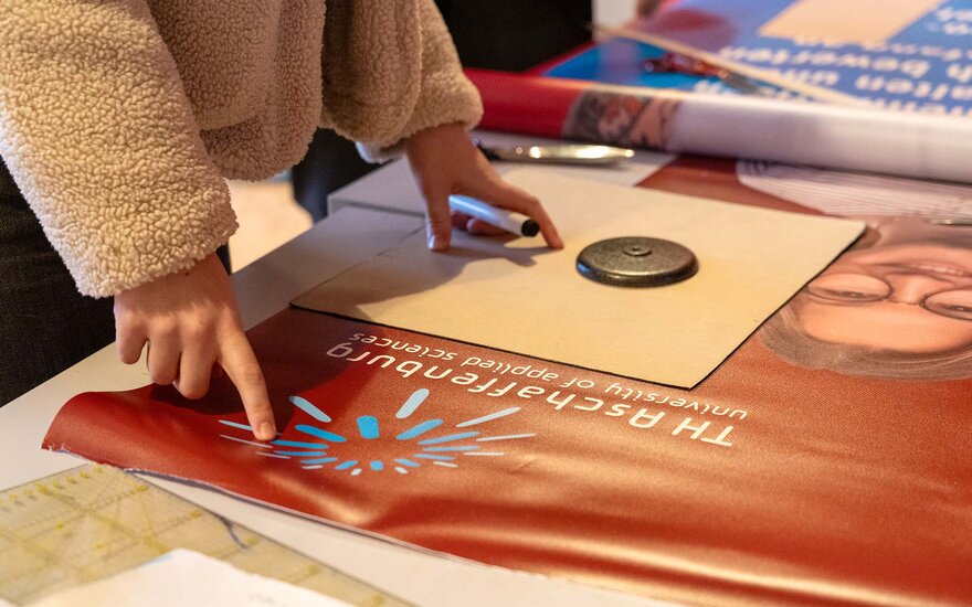 Ein altes, lilarotes Plakat der TH Aschaffenburg befindet sich auf einer Werkbank. Ein anonymes Mitglied des Gesta e.V. legt ein Schnittmuster auf das Plakat und streicht es glatt.