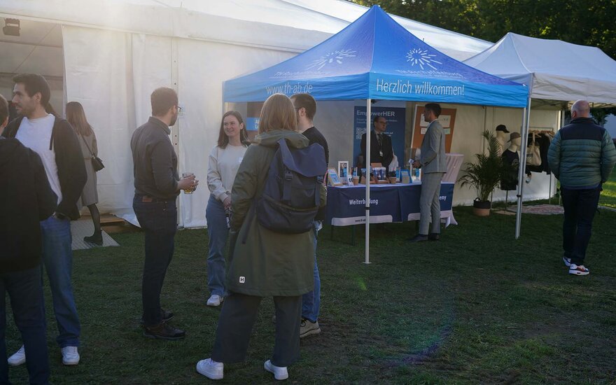 Personen in kleinen Gruppen stehen auf der Campuswiese vor dem Zelt und einem Infostand mit blauem TH-Pavillon