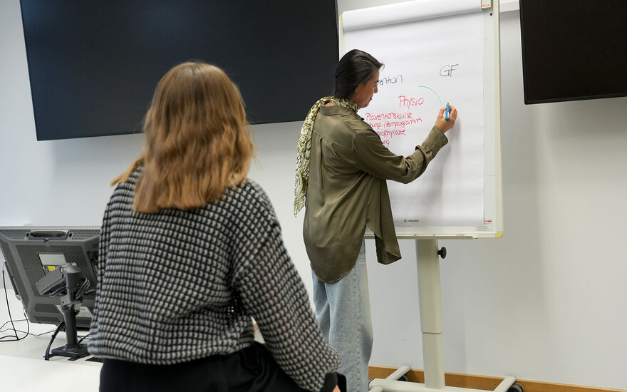 Eine Studentin skizziert Themen aus ihrem Berufsalltag an einem Whiteboard, während eine andere Studentin ihr zuhört