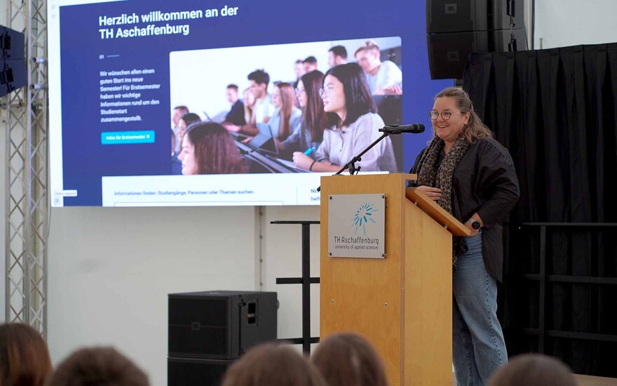 Frau mit braunen Locken, Brille und dunkler Jacke am Rednerpult vor Leinwand