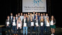 23 Studierende stehen in mehreren Reihen hintereinander mit ihren Zeugnissen in der Hand auf der Bühne vor einem Banner mit dem TH-Logo
