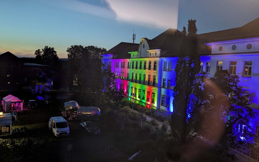 in Regenbogenfarben beleuchtete Fassade eines weißen Hochschulgebäudes in der Dämmerung