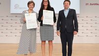 Zwei Hochschulvertreterinnen stehen mit Zertifikat in der Hand neben dem Geschäftsführer vor Logowand