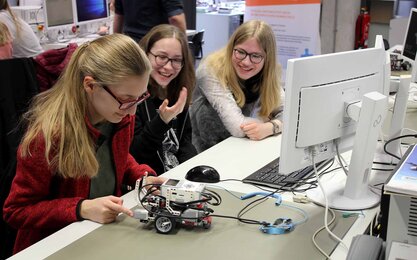 Schüler*innen sitzen an einem Schreibtisch und beschäftigen sich mit einem kleinen Roboter. 
