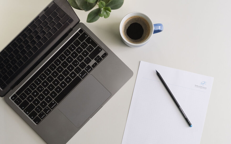 Auf einem Tisch steht ein Laptop, ein Kaffee, eine Pflanze und Papier mit Stift.