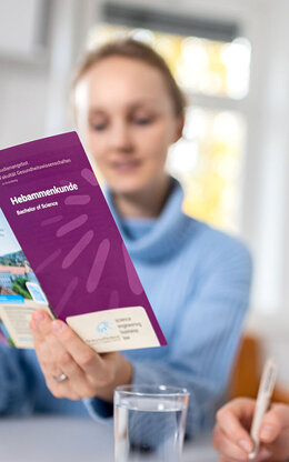 Eine Mitarbeiterin zeigt einer Studentin einen lilanen Flyer mit der Aufschrift "Hebammenkunde".