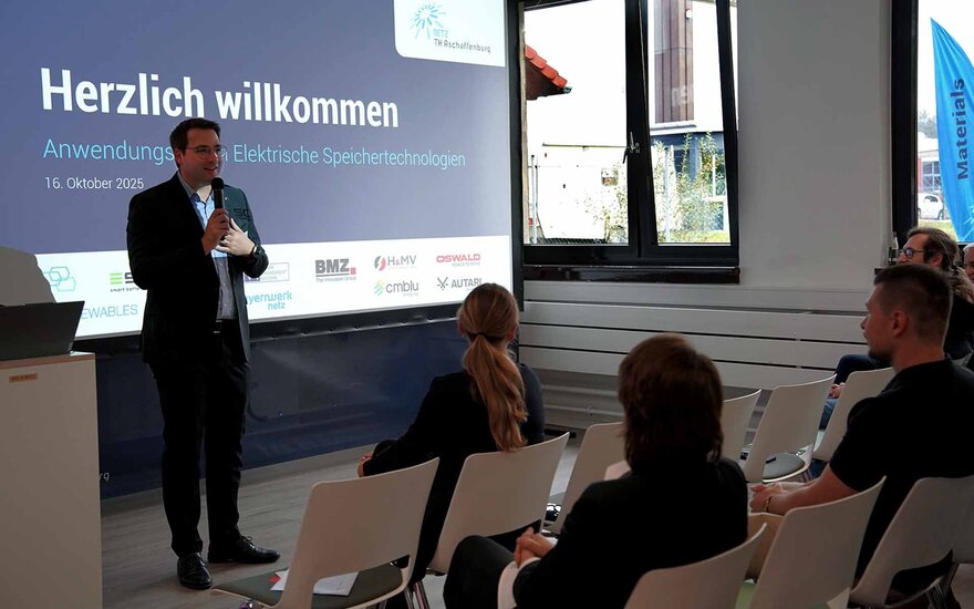 Ein Mann im Anzug hält eine Präsentation. Hinter ihm ist ein Bildschirm mit Grußworten.