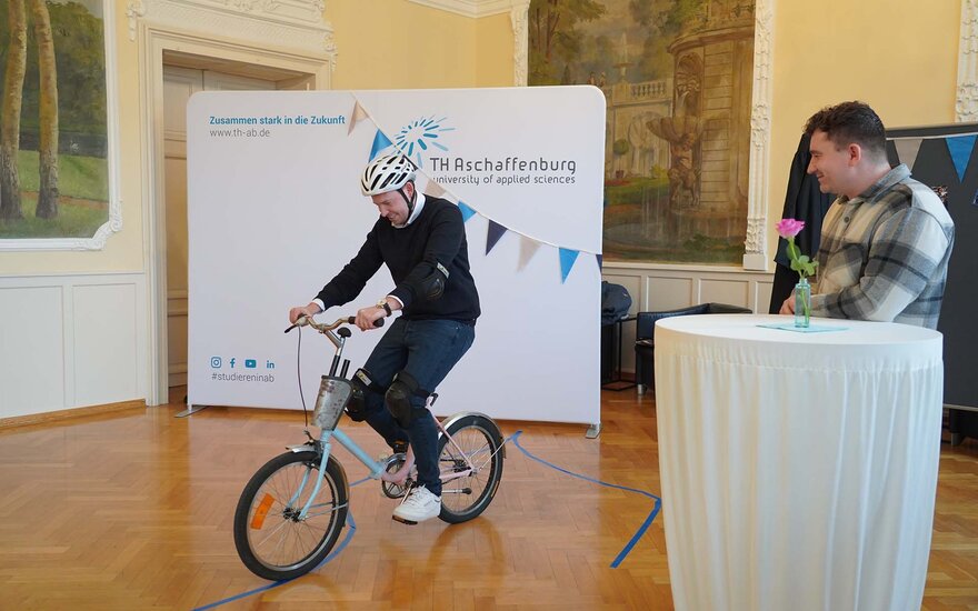 Timm fährt mit Helm auf einem kleinen Fahrrad, am Stehtisch neben ihm der lächelnde Manuel 