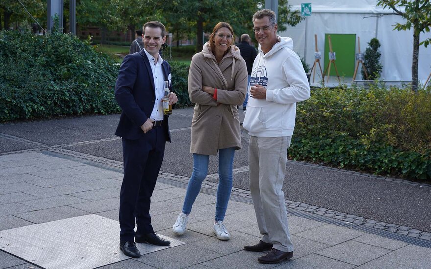 Ein junger Mann mit dunklem Anzug und weißem Hemd, eine junge Frau mit langen braunen Haaren, Brille, beiger Jacke und Jeans und ein Mann mittleren Alters mit dunklen Haaren, Brille, weißem TH-Hoodie und beiger Hose stehen nebeneinander vor dem Zelt und lachen