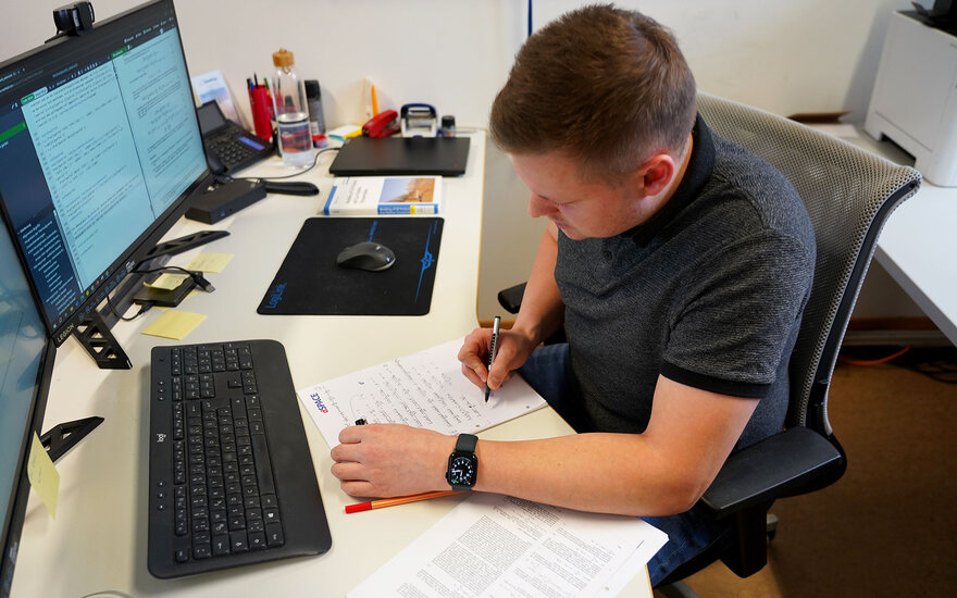 Luca Spielmann sitzt an seinem Arbeitzplatz und schreibt etwas in einem Notizblock während vor ihm auf dem Tisch noch ein Computer zu sehen ist.