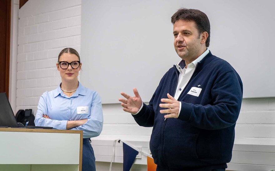 Schwarzhaariger Mann mit dunkler Jacke spricht stehend vor weißer Leinwand neben Frau mit Brille