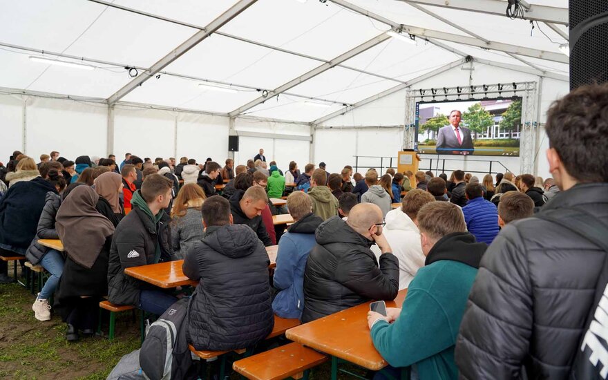 Studierende sitzen in einem Zelt auf Bänken vor einer Videoleinwand