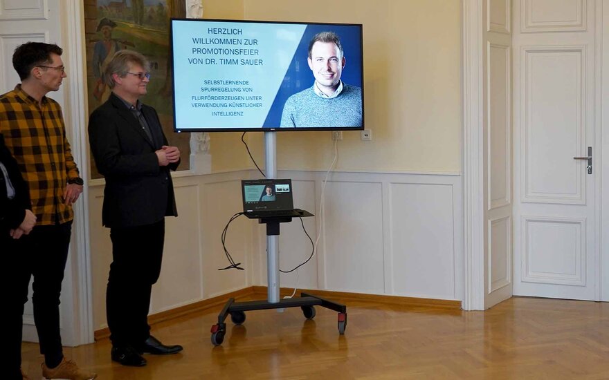 Monitor mit Foto von Timm und Promotionsthema, zwei Männer stehen links daneben