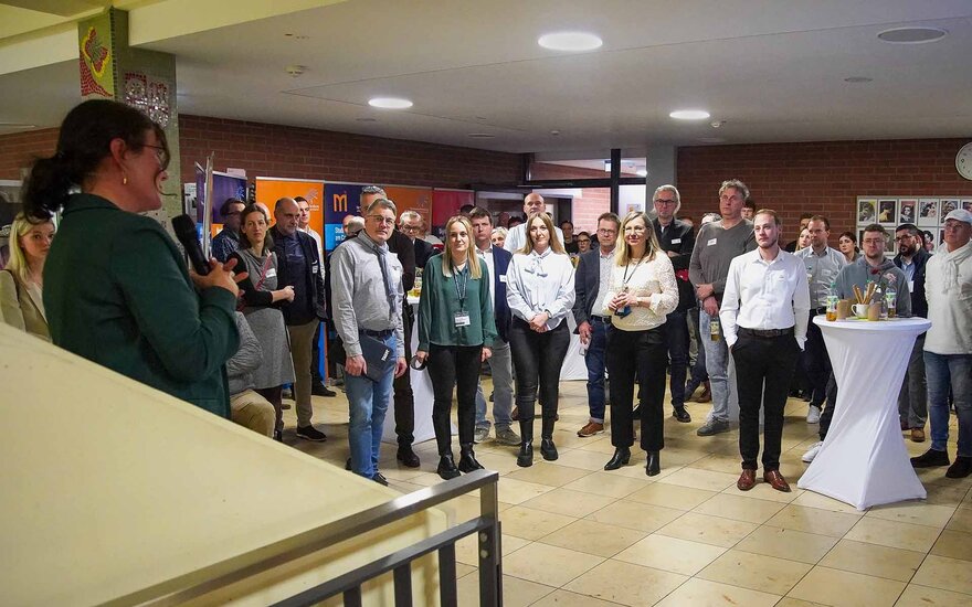 Große Gruppe von Teilnehmenden steht im Foyer und schaut zu vortragender Frau mit Mikrophon