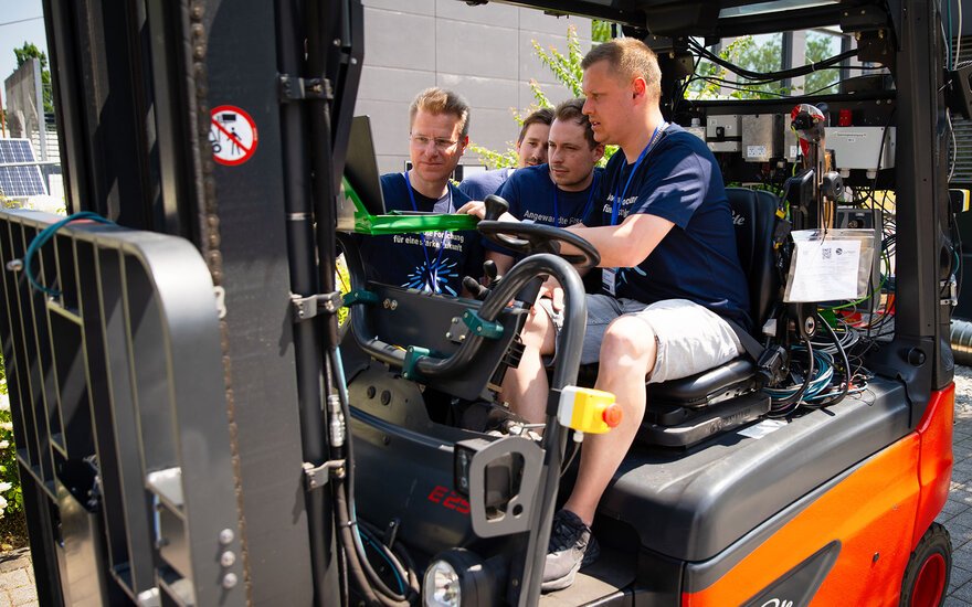 Luca Spielmann sitzt in einem Gabelstapler während drei andere Männer mit ihm zusammen auf einem Laptop schauen.