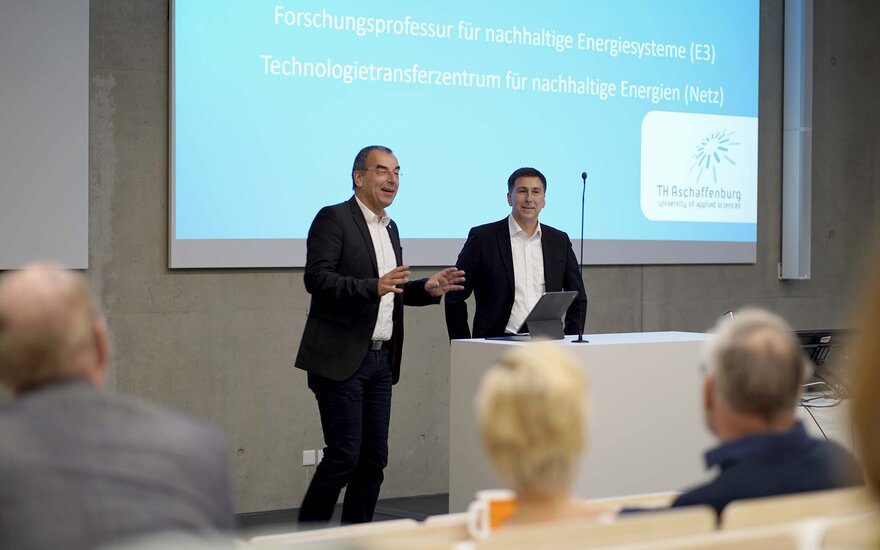 Zwei Männer in schwarzen Anzügen stehen vor der blauen Beamerleinwand im Hörsaal