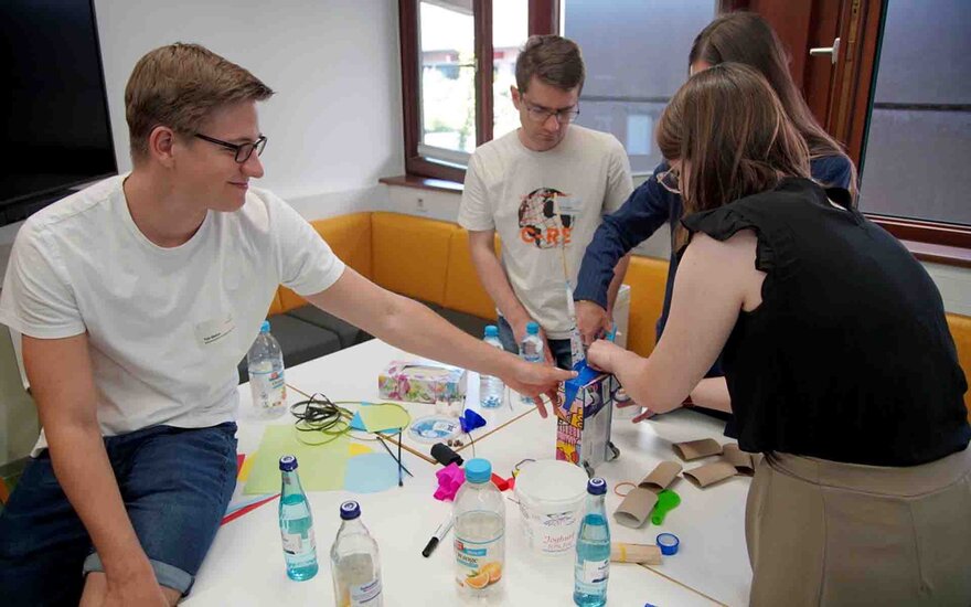 Studierende und Unternehmensvertreter bauen aus leeren Flaschen am Tisch ein Auto