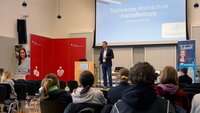 Ein Professor der Fakultät Wirtschaft und Recht steht in der Aula neben dem Rednerpult und spricht zu den sitzenden Schülerinnen und Schülern. Im Hintergrund ist auf einer blauen Leindwand der Schriftzug der TH Aschaffenburg zu sehen. Daneben steht einer rote Wand der Sparkasse.