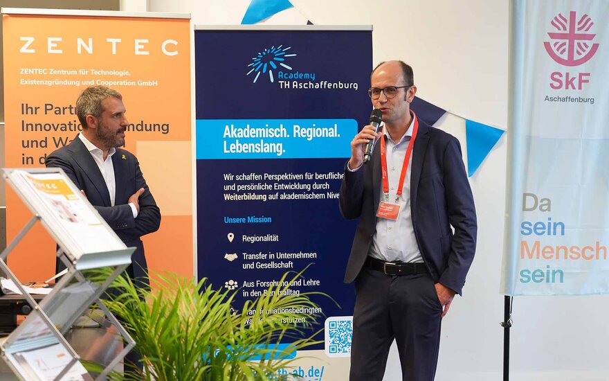 Mann mit Mikrofon in der Hand. Daneben steht ein Mann mit verschränkten Armen.