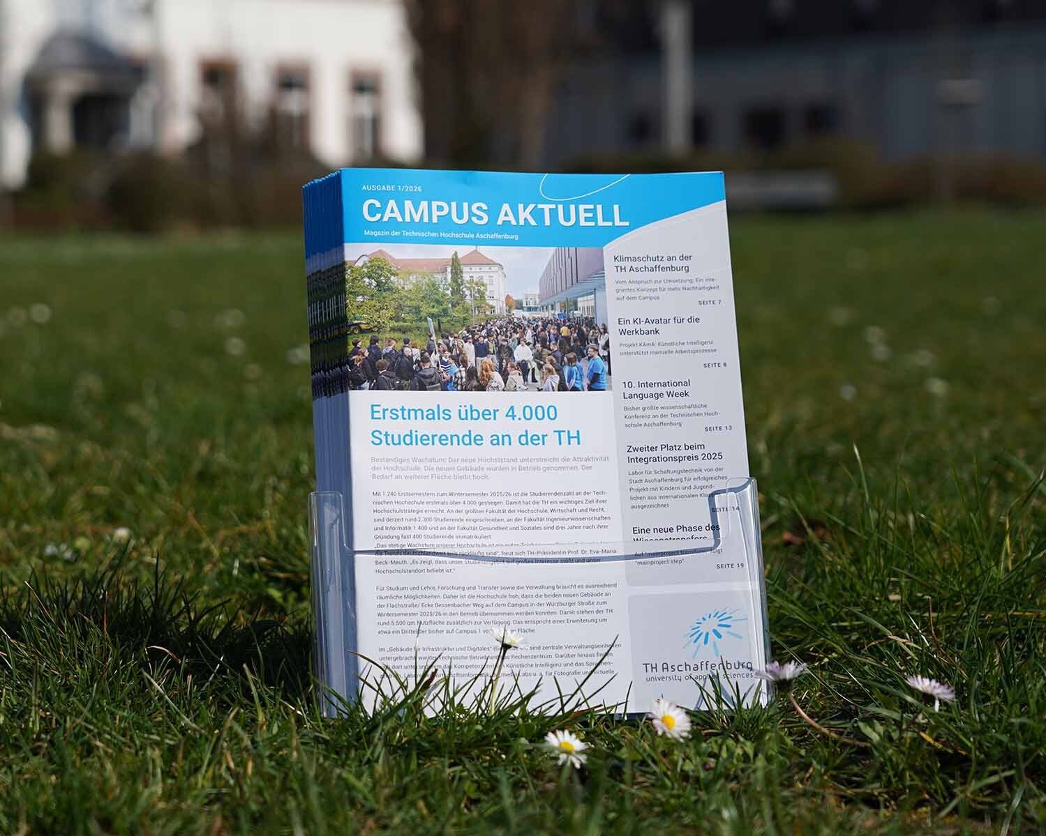 ochschulmagazine im Prospektständer auf Wiese mit Gänseblümchen vor Gebäuden der TH