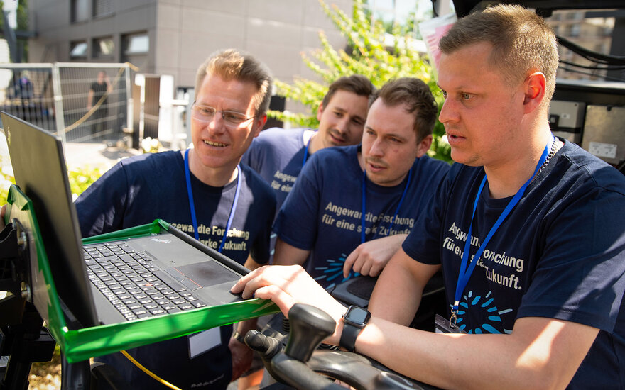 Nahaufnahe von Luca Spielmann, der in einem Gabelstapler sitzt während drei andere Männer mit ihm zusammen auf einem Laptop schauen. 