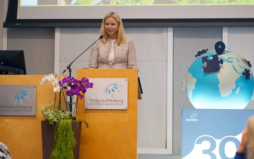 Blonde Frau in beigem Kostüm am Rednerpult mit TH-Logo und Keyvisual mit Weltkugel und 30-Jahre-Logo nebendran