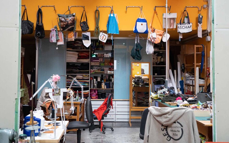 Nähwerkstatt von Gesta e.V. im Überblick. Zu sehen sind unterschiedliche Upcycling-Produkte, wie Taschen und Beuteln, die über einen mit Nähzubehör ausgestatteten Materialscrank aufgehängt sind. Im Vordergrund befinden sich Werkbänke mit Nähmaschinen und bereits angefangenen Projekten.