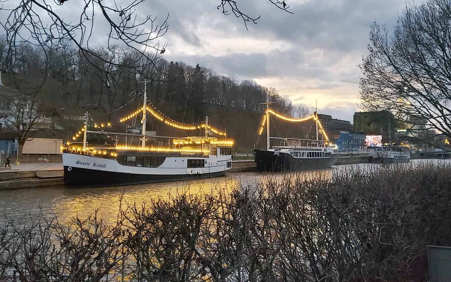 Zwei mit Lichterketten beleuchtete Schiffe am Flussufer bei wolkigem Himmel