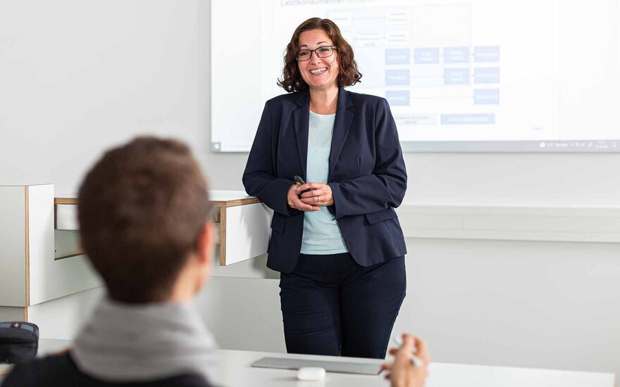Eine Frau im dunklen Blazer und dunkler Hose steht lächelnd am Rednerpult den Studierenden zugewandt