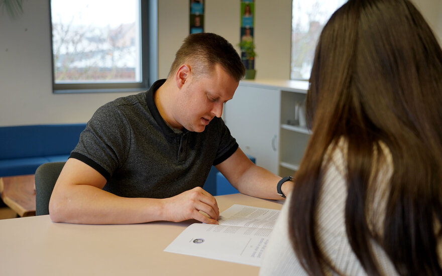 Luca Spielmann sitzt an einem Tisch während er mit einer Frau spricht und zeigt auf etwas in den Papieren, die vor ihm liegen.
