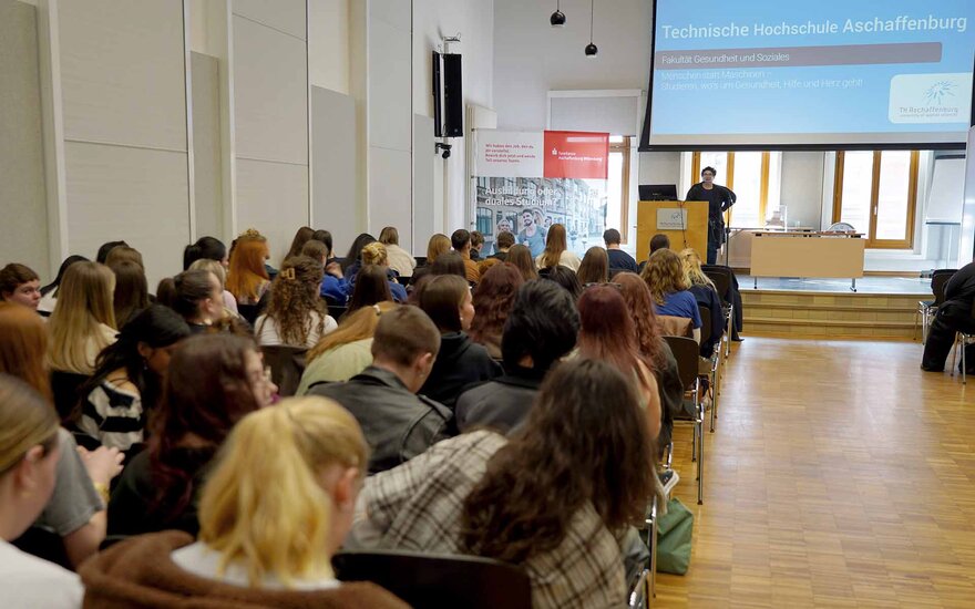 Mit sitzenden Schülerinnen und Schülern gefüllte Aula bei Vortrag mit Frau am Rednerpult