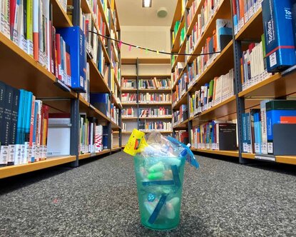 auf dem Boden vor mehreren Bücherregalen steht ein türkisfarbener Becher, der mit Ohrenstöpseln und einem Papierschirmchen gefüllt ist