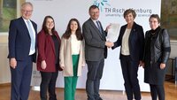 Gruppenbild mit zwei Männern und vier Frauen vor der TH-Logo-Wand im Hocksaal, Schulleiter und Präsidentin geben sich die Hand und halten den Vertrag