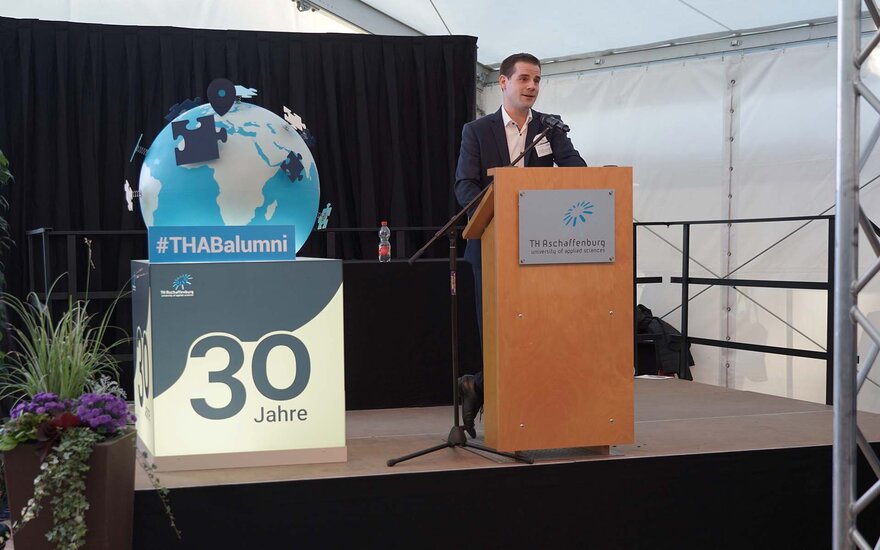 Junger Mann steht in dunklem Anzug an Rednerpult aus Holz mit TH-Logo darauf, links daneben steht ein beleuchteter großer Würfel mit einem blauen 30-Jahre-Schriftzug und einem blau-grauen Globus obendrauf