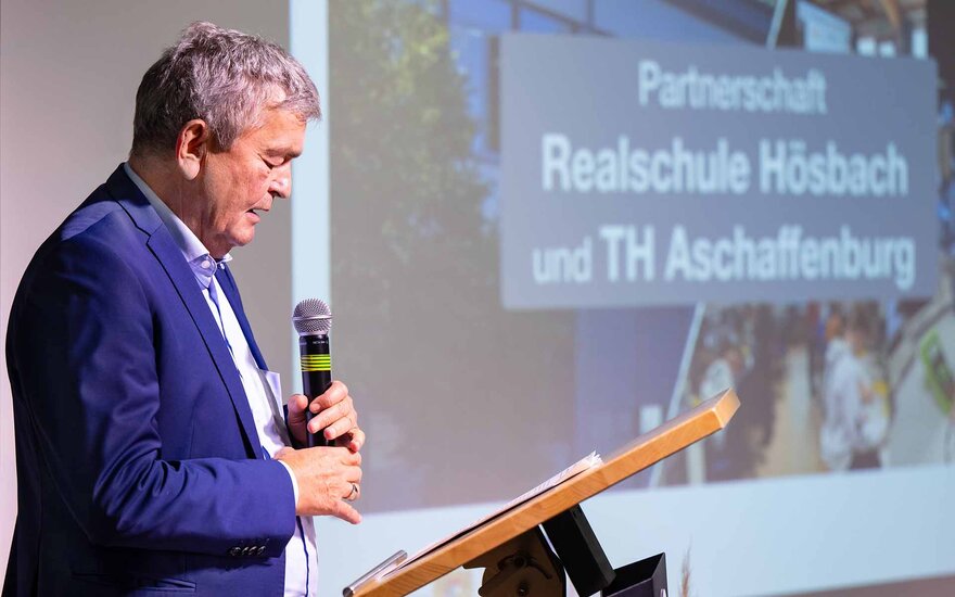 Grauhaariger Mann spricht mit Mikrophon am Rednerpult vor Leinwand mit Schriftzug zur Partnerschaft von RS und TH
