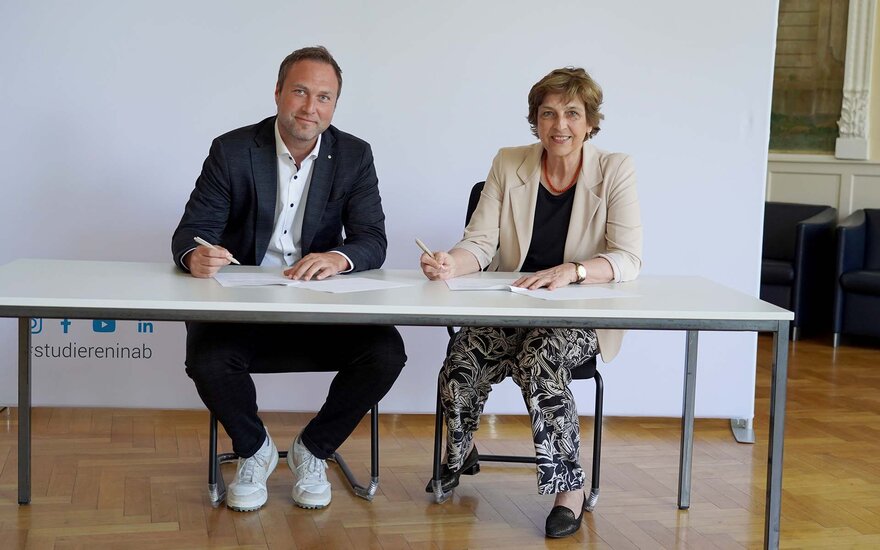 Caritas-Vorstand im dunklen Anzug und TH-Präsidentin im beigen Blazer unterzeichnen im Sitzen am Tisch Vertrag