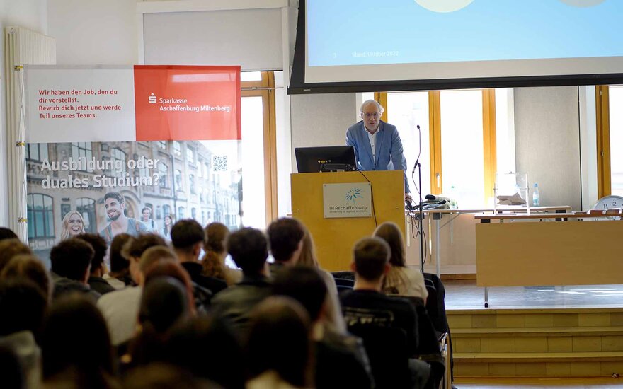 Professor spricht in der Aula am Rednerpult zu den Schülerinnen und Schülern