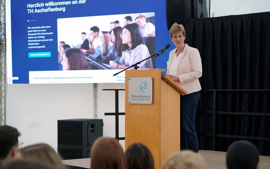 Frau mit blonden kurzen Haaren, Brille und rosa Blazer am Rednerpult vor Leinwand