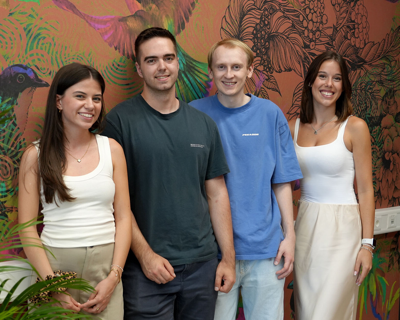 Vier Studierende, Christoph, Clarissa, Emilia und Marc, stehen lächelnd vor einer bunten Wand