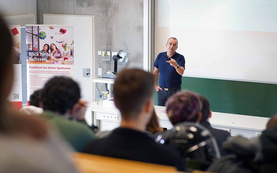 Professor steht vor Leinwand und spricht zu Schülerinnen und Schülern im ansteigenden Hörsaal
