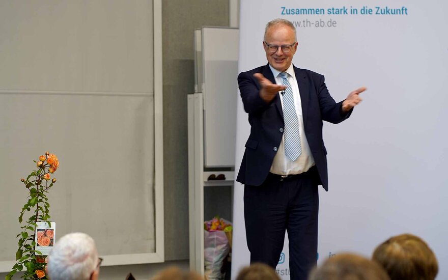 Mann mit grauen Haaren, Brille und schwarzem Anzug spricht mit einladender Geste zum Publikum