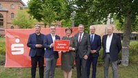 Präsidentin und Sparkassenvorstand mit rotem Schild mit Spendensumme in der Hand sowie Oberbürgermeister und drei Professoren in dunklen Anzügen stehen nebeneinander auf Campuswiese vor rotem Sparkassenbanner mit Backsteingebäude und Bäumen im Hintergrund