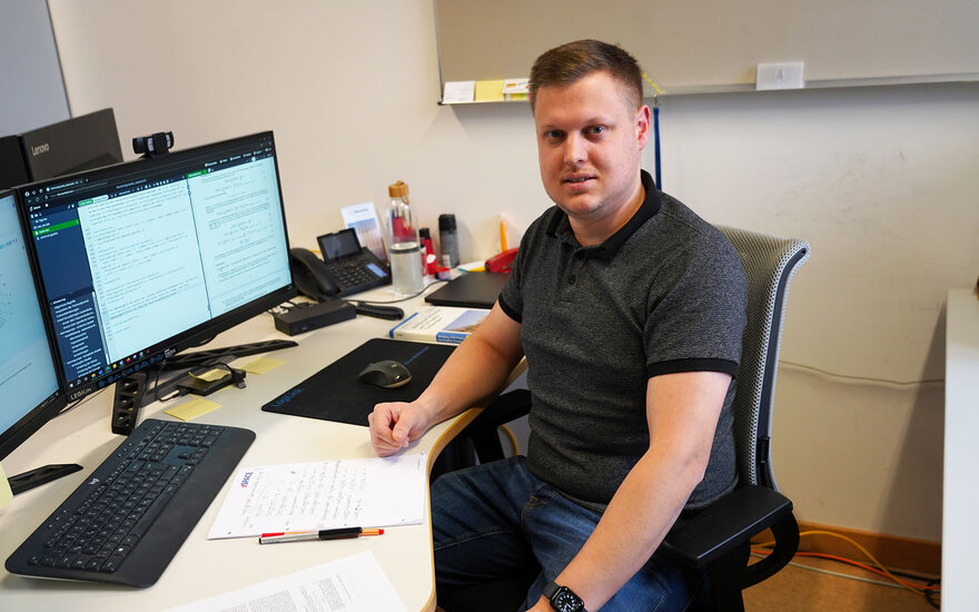 Luca Spielmann sitzt an seinem Arbeitzplatz, wo ein Computer und ein Notzblock zu sehen sind, und schaut direkt in die Kamera.