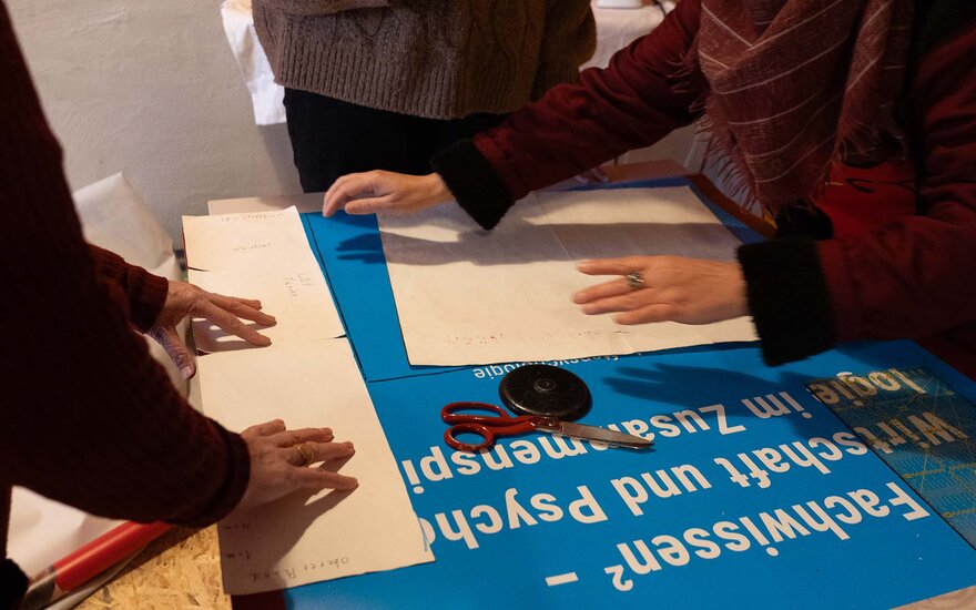 Auf einer Werkbank sieht man ein altes, blaues Banner der TH Aschaffenburg. Drei Mitglieder des Gesta e.V., von denen man nur Arme und Hände sieht, sind gerade dabei ein Schnittmuster auf das Banner zu zeichnen.