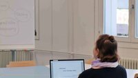 Eine Frau sitzt vor einem aufgeklappten Laptop mit Blick auf ein Flipchart