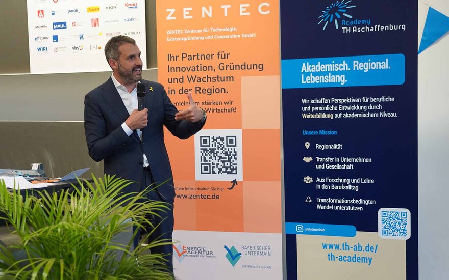 Sprechender Mann hält ein Mikrofon. Dahinter Roll-ups von Zentec und TH Academy