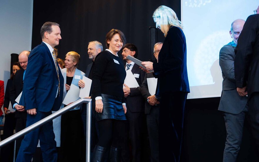 Eine blone Frau übergibt eine Urkunde an eine dunkelhaarige Fau. Daneben steht ein Mann im Anzug.