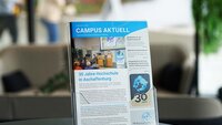 Hochschulmagazin im Prospektständer auf schwarzem Tisch im Foyer mit Sofa, Pflanze und vorbeilaufenden Personen