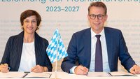 Die Hochschulpräsidentin sitzt links neben dem Wissenschaftsminister an einem Tisch und beide halten Stifte in der Hand, haben den Vertrag vor sich liegen und blicken in die Kamera
